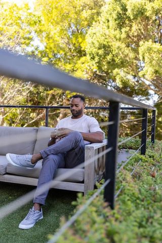 Mature african american man relaxing on sunlit terrace sofa using smartphone