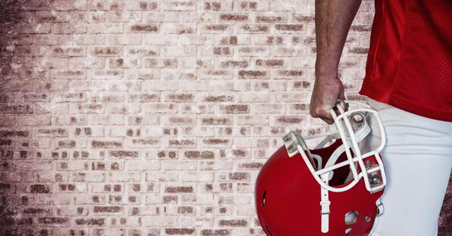 Football player stands against a brick wall, holding a red helmet in right hand. Dressed in sports uniform, including a red jersey and white pants. Ideal for use in sports-themed material, athlete advertisements, team promotions, or safety gear catalogs.
