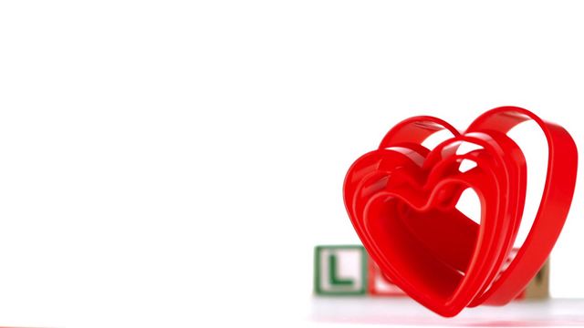 Bright red heart-shaped ornaments are gracefully falling in the foreground. In the blurred background, alphabet blocks spell out the word 'love', adding to the romantic and playful theme. This image can be used for expressing emotions, reinforcing themes of love and passion, or in Valentine's Day marketing materials.