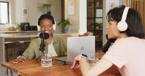 Diverse cohosts recording podcast at home table using microphones, laptop, headphones