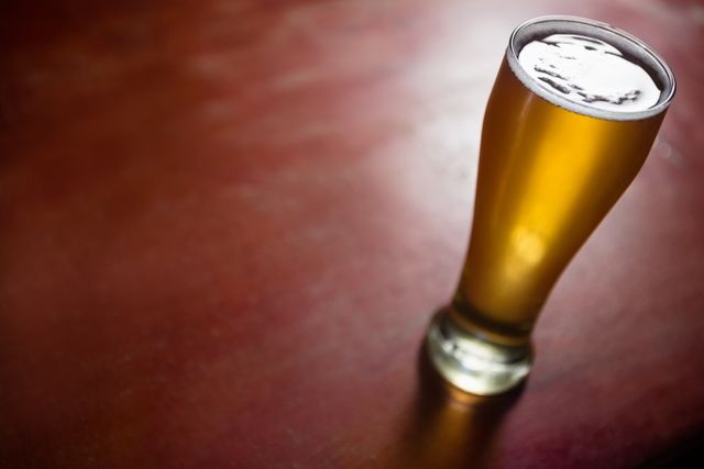 Close-up of a pilsner beer glass filled with golden beer, placed on a wooden table. Ideal for use in advertisements for bars, pubs, breweries, and beer brands. Can also be used in articles or blogs about beer, brewing, and leisure activities.