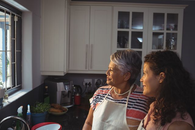 Mother and daughter looking through window in kitchen at home