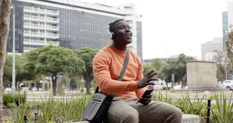 African American man listening to music and smiling holding smartphone on city bench