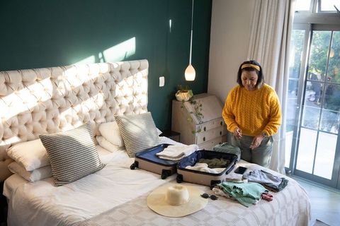 Mature woman packing suitcase in sunlit bedroom with straw hat and mustard sweater