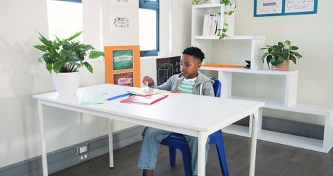 Young African American boy studying at white desk in bright classroom with notebooks and pen