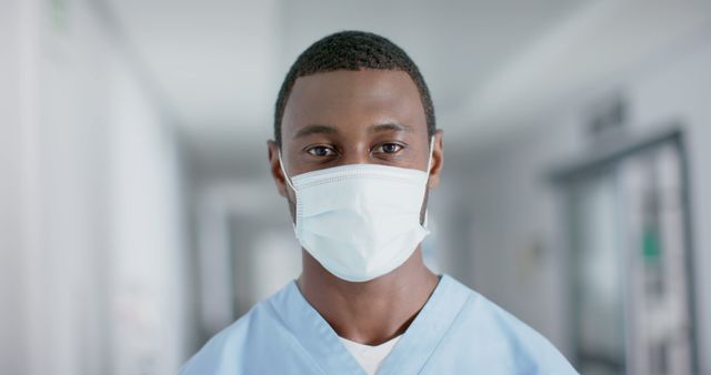 Portrait of happy african american male doctor wearing face mask in corridor at hospital. Medicine, healthcare, hygiene and hospital, unaltered.