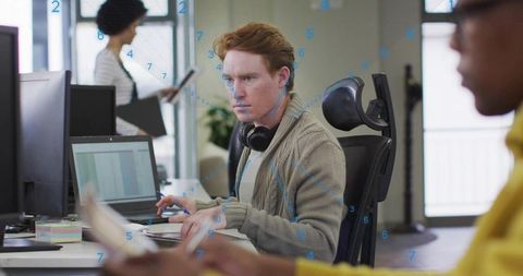 Focused redheaded man typing on laptop in open-plan office with headphones and paperwork