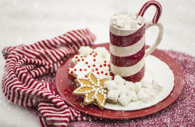 Close-up view of a mug filled with hot chocolate and topped with marshmallows, next to festive cookies on a red plate. The candy cane and striped red and white cloth add a touch of holiday cheer. Perfect for use in holiday marketing, festive cooking blogs, or social media promotions during the Christmas season.
