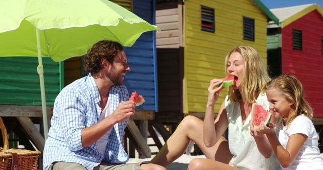 A Caucasian family enjoys slices of watermelon on a sunny beach, with colorful beach huts in the background. Capturing a moment of joy, the image reflects a relaxed summer atmosphere and family bonding time.