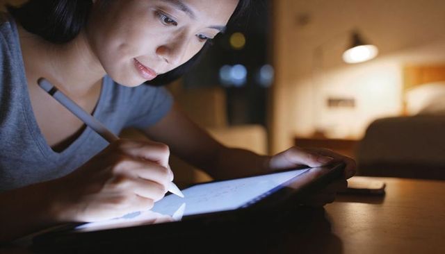 Late-night digital drawing by focused asian woman using stylus on tablet in cozy bedroom
