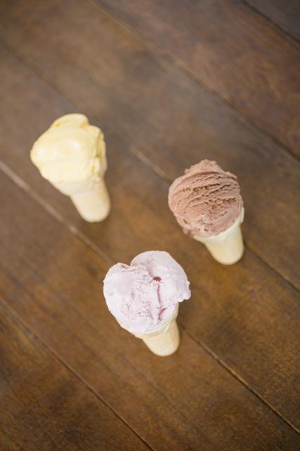 Three ice cream cones with vanilla, strawberry, and chocolate scoops placed on a wooden table. Ideal for use in food blogs, dessert menus, summer-themed promotions, and advertisements for ice cream shops.