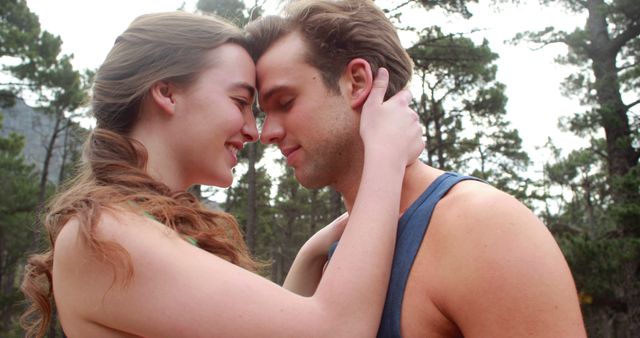 A young Caucasian couple shares an intimate moment, embracing and touching foreheads affectionately in a natural outdoor setting. Their close connection and the serene environment suggest a romantic and peaceful relationship.