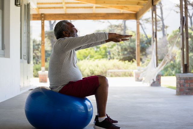 Senior man sitting on an exercise ball in a porch area, extending his arms forward while focusing on his fitness routine. Ideal for illustrating home workout, healthy aging, and active senior lifestyles in promotional materials, health blogs, or fitness guides.
