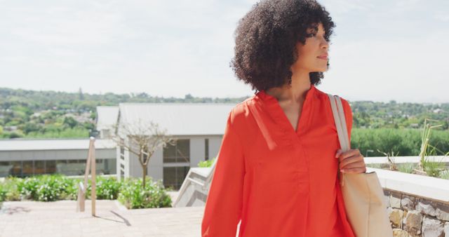 Woman with afro hairstyle walking outdoors in an urban environment. She wears a red blouse and carries a tote bag. There are buildings and greenery in the background on a sunny day. Ideal for use in advertisements, lifestyle blogs, fashion articles, or urban life-related content.