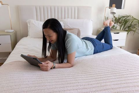 Mature asian woman lying on bed using tablet in minimalist neutral cozy bedroom relaxing