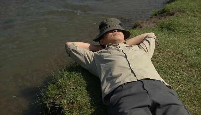 Man lying by lakeside wearing bucket hat and beige shirt relaxing on grassy shoreline under sun