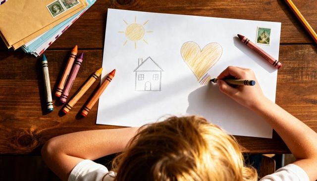 Child drawing heart and house with crayons on wooden table in sunlit cozy home moment