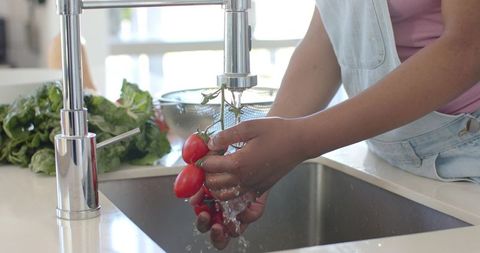 Washing vine tomatoes under chrome faucet in bright modern kitchen with denim apron