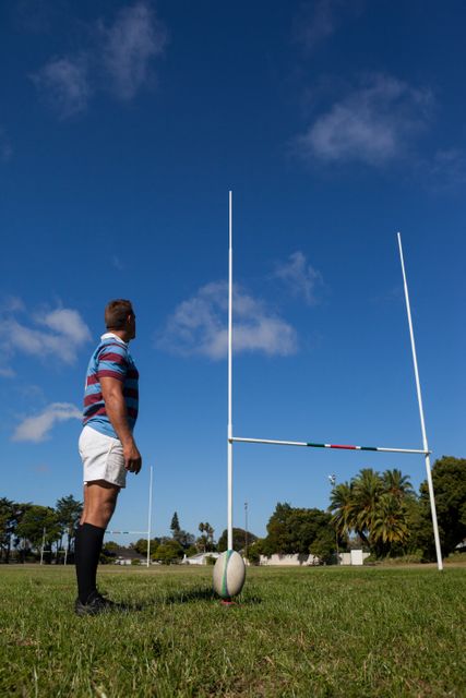 Rugby player standing on field looking at goal post under clear blue sky. Ideal for sports-related content, athletic training materials, motivational posters, and articles about rugby or outdoor activities.