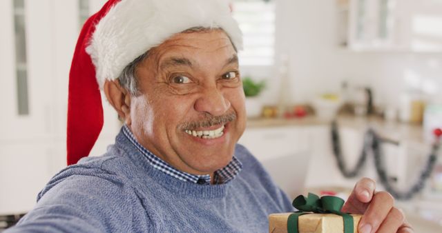 Image of happy senior biracial man in santa hat making christmas image call holding gift, laughing. Christmas, tradition, global communication, inclusivity and senior lifestyle concept.