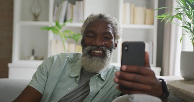 Senior man smiles while using his smartphone in a cozy living room. He sits on a couch wearing a casual green shirt. This image can be used for technology, communication, and elderly lifestyle concepts, promoting seniors staying connected and enjoying modern tech.