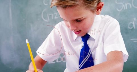 Focused schoolboy writing math problems at classroom desk with blue tie and chalkboard