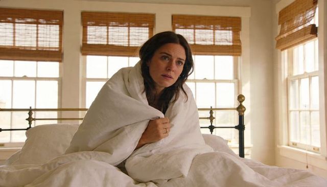 Woman sitting wrapped in white duvet gazing contemplatively in sunlit bedroom with bamboo blinds