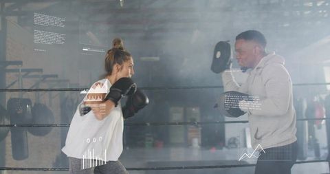 Female boxer training with coach holding mitts in moody warehouse gym with data overlays