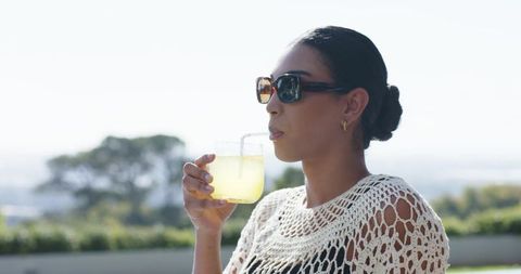 African american woman sipping lemonade on sunny terrace wearing crochet top and sunglasses