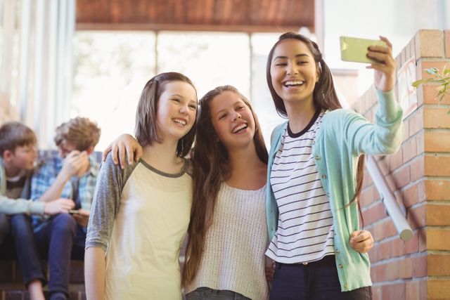 Three schoolgirls are taking a selfie with a mobile phone in a school corridor. They are smiling and appear to be enjoying their time together. This image can be used to depict themes of friendship, youth, and the use of technology among teenagers. It is suitable for educational materials, social media campaigns, and advertisements targeting young audiences.