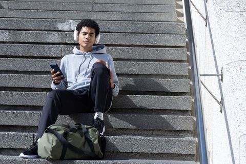 African american teen sitting on stairs listening to music holding phone and football