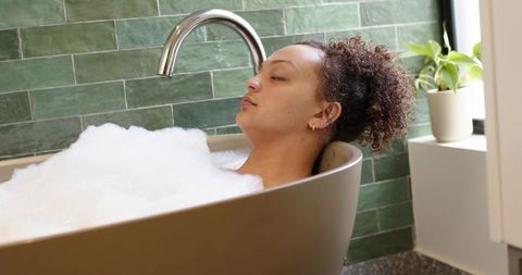 Relaxing woman soaking in freestanding bathtub with foam by green tiled wall, serene spa