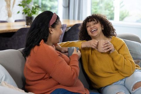 Laughing african american friends sharing coffee on cozy sofa, casual home conversation