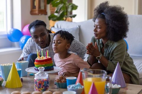 Family celebrating child birthday blowing candles on rainbow cake at home party