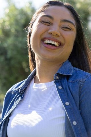 Laughing young woman wearing denim shirt and white ribbed top outdoors with sunlit bokeh