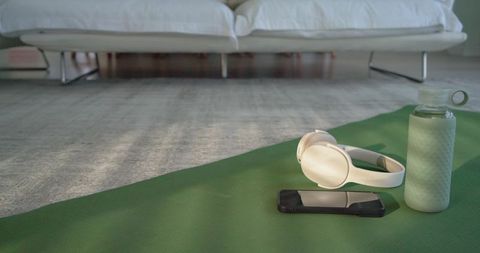 White wireless headphones and water bottle on green yoga mat near sofa minimalist wellness