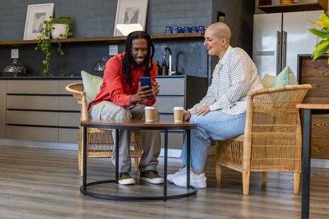 Sharing smartphone in modern kitchen lounge, diverse coworkers smiling over coffee cups