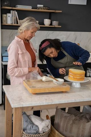 Intergenerational baking lesson: women decorating layer cake together on kitchen island