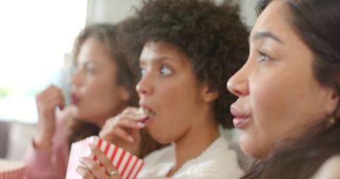Diverse women watching movie night on sofa eating popcorn and enjoying cozy home moment