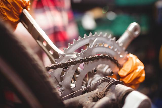 Mechanic wearing orange gloves repairing bicycle chain and gears in workshop. Ideal for content related to bike maintenance, mechanical work, cycling, repair services, and professional workshops.