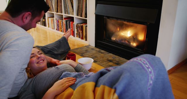 Couple lying together on floor near lit fireplace, covered with blankets, and smiling, enjoying cozy, warm evening at home. Bookshelves and cups enhance relaxed atmosphere. Perfect for content related to comfort, home living, winter moments, and relationships.