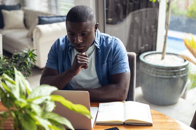 African American man working from home, looking at laptop with hand on chin. Ideal for illustrating remote work, home office setups, business, technology use, and concentration. Suitable for articles on productivity, work-life balance, and modern work environments.