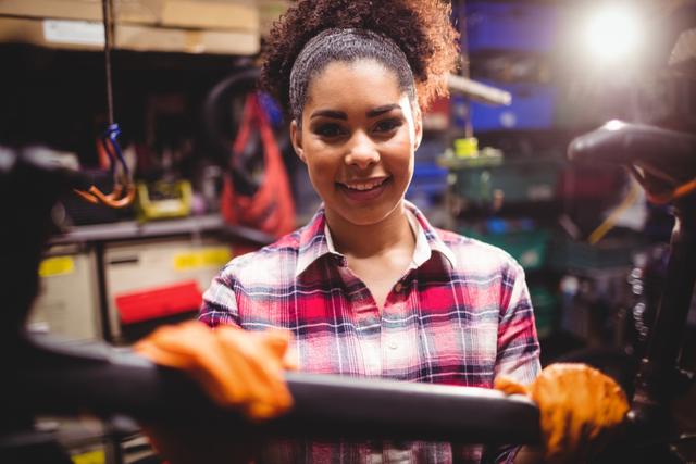 Image depicts a confident female mechanic holding a bicycle in a workshop. She is wearing a plaid shirt and orange gloves, with various tools and equipment in the background. This image is ideal for promoting services related to bicycle repair, showcasing female professionals in mechanical fields, or illustrating concepts of skilled trades and technical work.