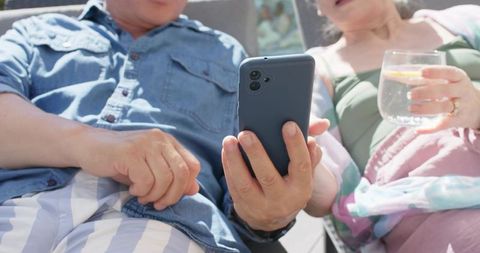 Mature diverse couple lounging poolside sharing smartphone while sipping lemon water