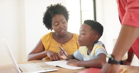 African American Family Guiding Son with Homework Using Laptop at Sunlit Wooden Table