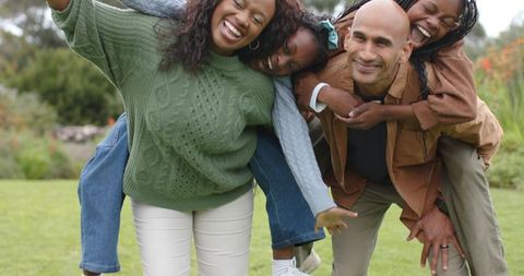 Diverse family laughing while parents give piggyback rides in sunny garden park