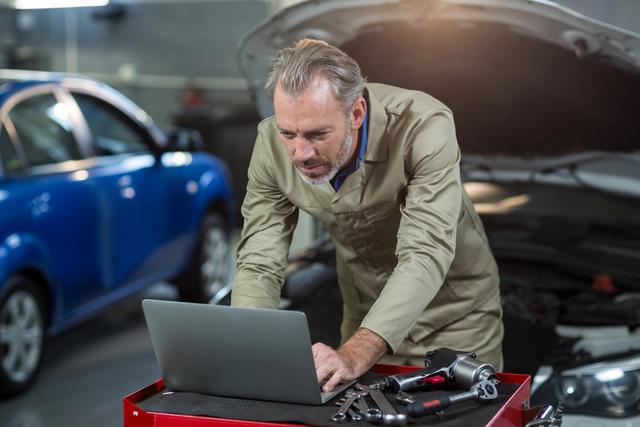 Mechanic using a laptop while working in a car repair garage. This image is useful for illustrating the integration of technology in automotive diagnostics and repair. Ideal for content related to modern car repair techniques, automotive industry advancements, or training and educational materials for mechanics.