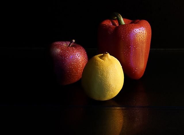 Red apple, yellow lemon, and red bell pepper arranged on a black reflective surface with water droplets enhancing the freshness. Ideal for use in healthy eating, nutrition, or kitchen-related content.