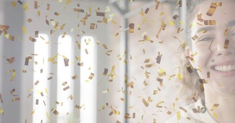Smiling woman celebrating with falling gold confetti backlit by tall windows portrait