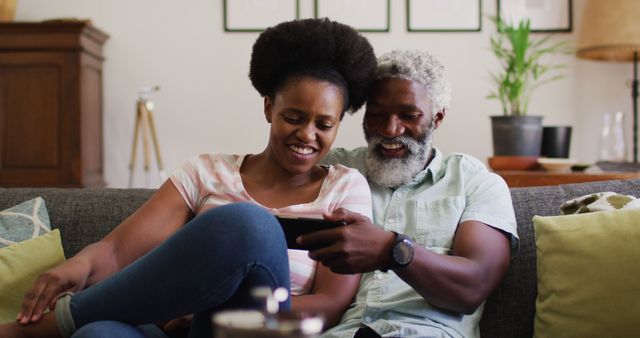 Elderly couple relaxing on a couch at home, watching a tablet together and smiling. Ideal for use in marketing materials focused on family bonding, senior living, and technology use among senior citizens, or for articles about modern elderly lifestyles, love, and togetherness.